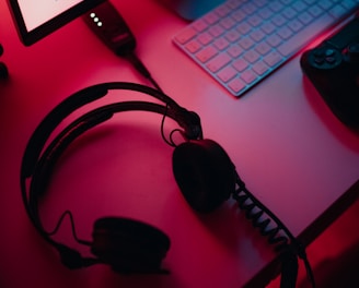 A dimly lit gaming setup with a monitor displaying a game's menu on the screen. A mechanical keyboard sits in front of the monitor. A pair of over-ear headphones and a game controller rest on the desk. The scene is bathed in a red and blue light, creating a moody gaming atmosphere.