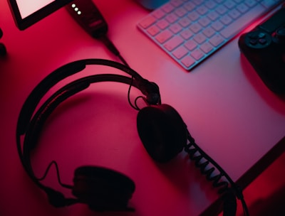 A dimly lit gaming setup with a monitor displaying a game's menu on the screen. A mechanical keyboard sits in front of the monitor. A pair of over-ear headphones and a game controller rest on the desk. The scene is bathed in a red and blue light, creating a moody gaming atmosphere.