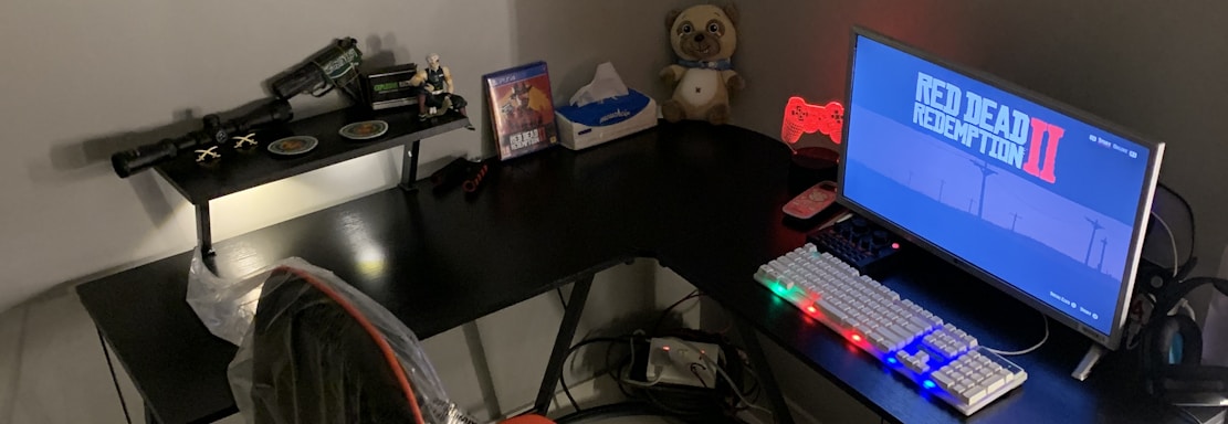 A gaming setup features a large corner desk with a computer monitor displaying the game Red Dead Redemption II. The desk holds a gaming chair still wrapped in protective plastic, a gaming keyboard with colorful backlighting, a game controller, and decorative items like a plush toy. A blanket and a pillow are on the side, suggesting a cozy setup.