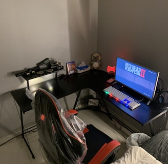 A gaming setup features a large corner desk with a computer monitor displaying the game Red Dead Redemption II. The desk holds a gaming chair still wrapped in protective plastic, a gaming keyboard with colorful backlighting, a game controller, and decorative items like a plush toy. A blanket and a pillow are on the side, suggesting a cozy setup.