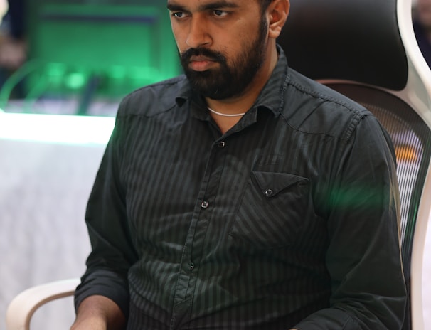 A person is seated in a chair, focused intently on something, with hands positioned on a gaming console or arcade controller. The setup appears to be indoors, with visible screens and lighting in the background, suggesting an electronic gaming environment.